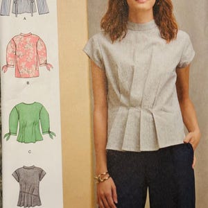 Puede incluir: Una mujer con un top gris claro de manga corta con la parte delantera plisada y cuello alto, combinado con pantalones azul oscuro. La imagen también muestra cuatro diseños de blusas diferentes: a rayas, floral, verde y gris.