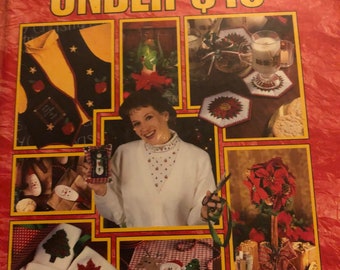 May include: A book titled "Christmas Gifts Under $10" with a red cover and a collage of images of various Christmas crafts. The crafts include a snowman, a poinsettia, a mug cozy, and a Christmas stocking.