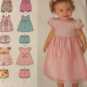 May include: A Simplicity sewing pattern, 1701A, with illustrations of baby dresses and bloomers. A baby girl wears a pink dress with ribbon and rose accents, and matching shoes. The pattern includes sizes XXS-L.