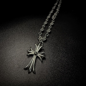 Könnte beinhalten: Ein silberfarbener Metallkreuz-Anhänger an einer passenden Kette. Das Kreuz hat ein ausgestelltes, verziertes Design mit einem zentralen kreisförmigen Detail. Die Kette besteht aus miteinander verbundenen runden Gliedern mit einem Muster.