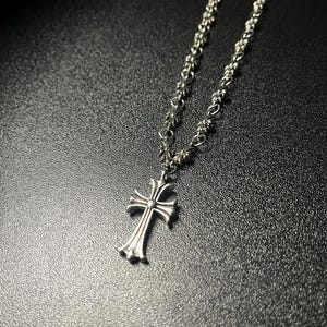 Könnte beinhalten: Eine silberfarbene Metallhalskette mit einem Kreuzanhänger, der ausgestellte Enden und ein zentrales kreisförmiges Detail aufweist. Die Kette besteht aus kleinen, sich wiederholenden dekorativen Gliedern, die zum Design des Kreuzes passen.