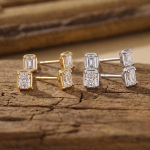 May include: Two pairs of stud earrings featuring emerald-cut clear stones. One pair is set in yellow gold-toned metal, and the other pair is set in silver-toned metal. Each earring has a post back and a rectangular stone held by four prongs.