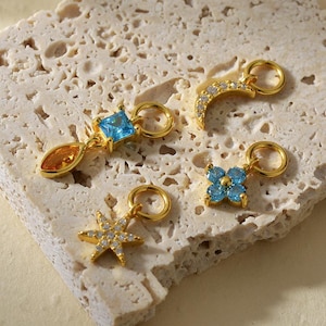 May include: Four gold-toned metal charms featuring various shapes and colorful gemstones. One charm is a square blue stone with a teardrop orange stone, one is a crescent moon with clear stones, one is a star with clear stones, and one is a blue flower.