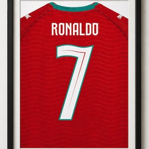 Puede incluir: Impresión enmarcada de una camiseta de fútbol roja con el nombre "RONALDO" y el número "7" en blanco. La camiseta tiene un cuello verde y ribetes blancos en el número. La impresión está en un marco negro.