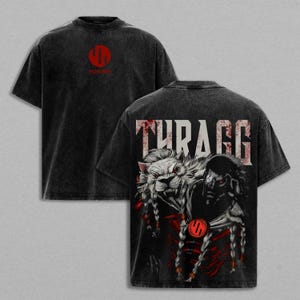 May include: Black t-shirt with a distressed look. The front features a red logo, while the back displays the word "THURAGG" above a graphic of a lion-like creature and a warrior with red accents. The shirt has short sleeves.