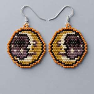 May include: A pair of circular beaded earrings featuring a crescent moon design. The beads are arranged in a grid pattern using colors of orange, black, cream, and muted purple. Each earring hangs from a silver-toned metal hook for attachment.