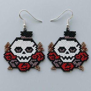 May include: A pair of beaded earrings featuring a skull wearing a black top hat, surrounded by red and brown floral elements. The skull is white with black details, and the earrings hang from silver-toned metal hooks against a plain background.