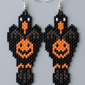 May include: A pair of dangle earrings crafted from small square beads in black, orange, and white. The earrings feature a bird-like shape with an orange circular pattern on the chest, orange beaks, and white eyes, suspended from silver-toned hooks.