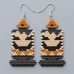 May include: A pair of beaded drop earrings shaped like a lit candle. The design features a cream-colored candle body with a black facial expression, a dark base, and a flame at the top rendered in orange, yellow, and black glass seed beads.
