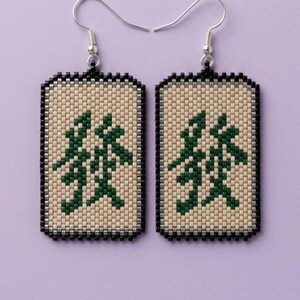 May include: A pair of rectangular beaded earrings featuring a cream-colored background with a dark green Chinese character symbol in the center. The edges are bordered by a row of silver beads and a final outer frame of black beads with silver hooks.