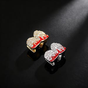 Può includere: Due set di grillz a forma di cuore. Un set è dorato con fiamme in smalto rosso e strass trasparenti. L'altro set è argentato con fiamme in smalto rosso e strass trasparenti. I grillz sono esposti su una superficie nera.