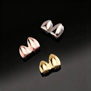 May include: Three sets of metallic tooth caps in silver, rose gold, and gold. Each set consists of two caps, designed to fit over the front teeth. The caps have a smooth, polished finish and are displayed against a black background.