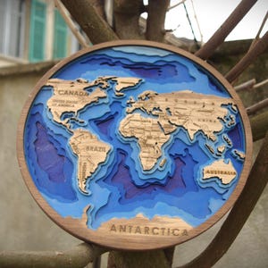 Può includere: Una mappa del mondo rotonda in legno con un design a strati. I continenti sono marrone chiaro e gli oceani sono blu. La parola "ANTARCTICA" è in basso. La mappa è montata su un ramo d'albero.