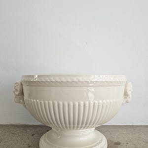 May include: A cream-colored, ceramic urn with a wide, open bowl shape. The urn features vertical ribbed detailing on the body, a rope-like design around the rim, and decorative handles. It stands on a pedestal base, suitable for decorative use.