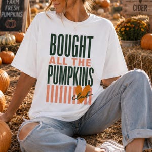 May include: A white short-sleeved t-shirt featuring the dark green and peach text BOUGHT ALL THE PUMPKINS CLUB with a small orange heart graphic and vertical peach stripes. The shirt is paired with light blue denim jeans with distressed knees.