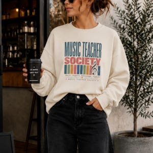 Puede incluir: Una sudadera de cuello redondo color crema con el texto MUSIC TEACHER SOCIETY en azul y rojo, un gráfico de barras verticales de colores, una clave de sol y el texto pequeño teach inspire harmonize impact MUSIC TEACHER SOCIETY.