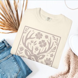 Peut inclure: T-shirt cr&egrave;me avec un motif de t&ecirc;te de cerf avec des bois, des fleurs et des feuilles dans un cadre rectangulaire. Le t-shirt est pr&eacute;sent&eacute; avec un jean bleu, une chaussure blanche et une &eacute;charpe beige. Le t-shirt porte une &eacute;tiquette "Comfort Colors".