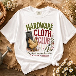 Puede incluir: Una camiseta blanca con un gráfico de una gallina marrón, un rollo de malla metálica y unas tijeras de podar. El texto dice HARDWARE CLOTH CLUB, We don't use chicken wire. We're not amateurs. El diseño incluye pequeñas flores y corazones.