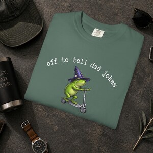 May include: A teal green sweatshirt with the text "off to tell dad jokes". The graphic shows a green frog wearing a wizard's hat and riding a scooter. The design is centered on the frog illustration.