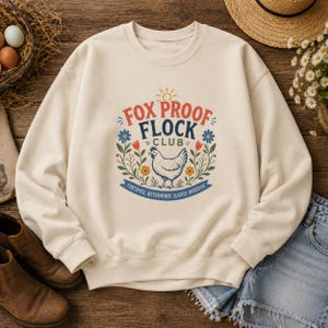 Può includere: Una felpa girocollo color crema con una grafica centrale raffigurante una gallina bianca, accenti floreali colorati e il testo "FOX PROOF FLOCK CLUB" in rosso e blu, con "FORTIFIED, DETERMINED, SLIGHTLY OBSESSIVE" stampato sotto.