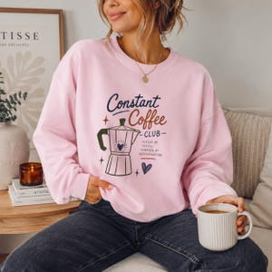 Puede incluir: Una sudadera de cuello redondo en color rosa claro con un gráfico de una cafetera italiana y el texto Constant Coffee CLUB, FUELED BY COFFEE, POWERED BY DETERMINATION, junto a pequeños iconos de corazones y estrellas en azul y marrón.