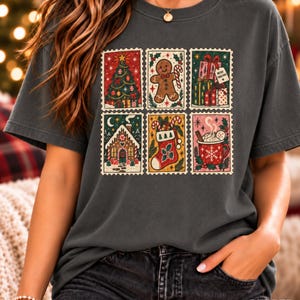 Puede incluir: Una camiseta gris carbón con un estampado gráfico de seis sellos postales rectangulares. Los sellos muestran ilustraciones festivas navideñas que incluyen un árbol de Navidad, un hombre de jengibre, regalos, una casa, un calcetín y una taza.