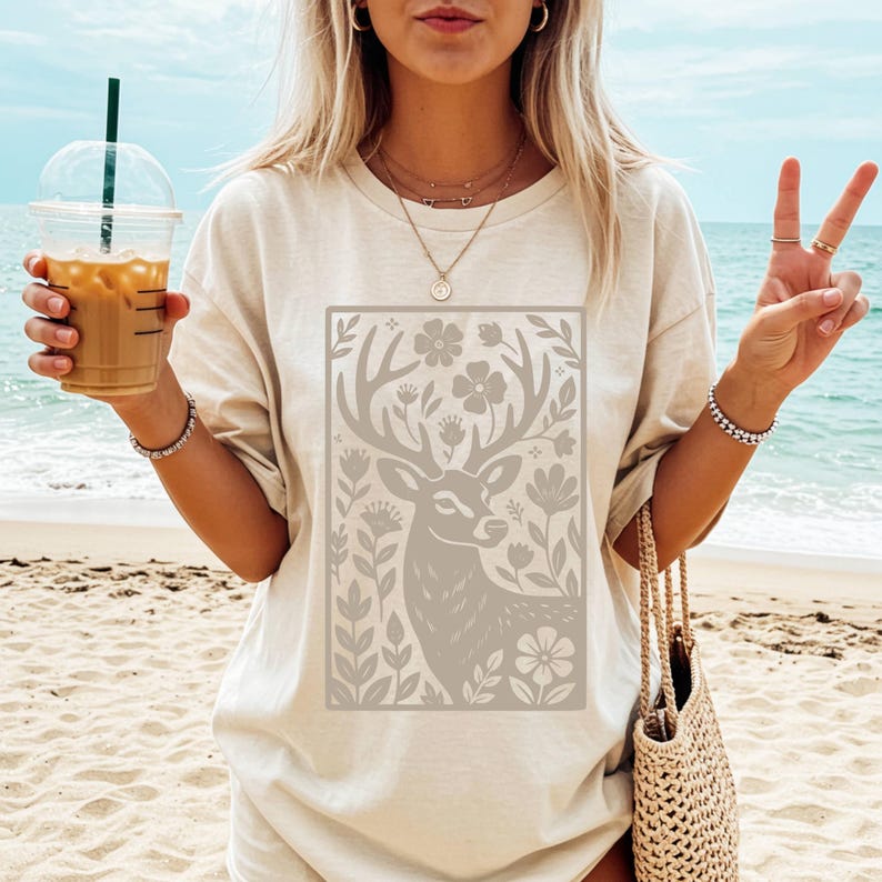Peut inclure: T-shirt beige avec un motif rectangulaire repr&eacute;sentant un cerf entour&eacute; de fleurs et de feuillages gris clair. Le t-shirt est port&eacute; sur une plage, avec un sac en paille.