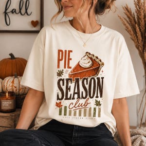 Op de afbeelding: Een crèmekleurig T-shirt met korte mouwen en een grafische print van een stuk pompoentaart met slagroom. De tekst luidt PIE SEASON club in bruine en oranje letters, vergezeld van kleine bladillustraties en verticale strepen.