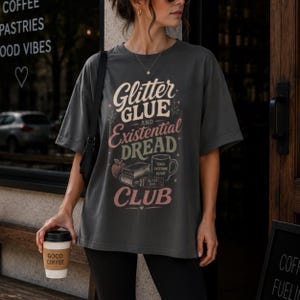 Könnte beinhalten: Ein anthrazitfarbenes T-Shirt mit dem Text Glitter Glue and Existential Dread Club in Creme, Rosa und Grün. Die Grafik zeigt einen Apfel, einen Bücherstapel und eine Tasse mit der Aufschrift Teach Overthink Repeat sowie eine Klebertube.