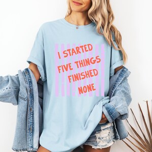 May include: Light blue t-shirt with the text "I STARTED FIVE THINGS FINISHED NONE" in orange and pink. The shirt has a relaxed fit and is paired with a denim jacket and shorts.
