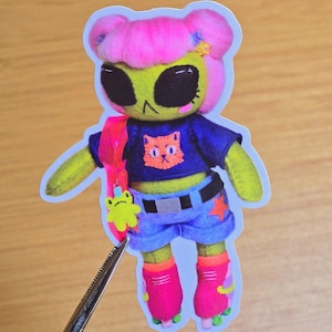 May include: A sticker of an alien doll with pink hair buns, a green face, and black eyes. The doll wears a blue shirt with an orange cat graphic, blue shorts, and pink roller skates. A pink backpack and a green frog charm complete the look.