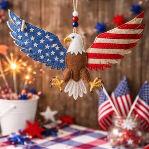 May include: A felt eagle ornament with outstretched wings, featuring the American flag design. The wings display blue with white stars and red and white stripes. The eagle has a white head, brown body, and yellow talons. The ornament hangs from a string with red, white, and blue beads.