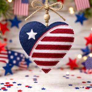 May include: A patriotic heart-shaped ornament, half blue with a white star and half red and white stripes, hangs from a twine bow. The ornament is part of a festive display with American flags and star-shaped confetti, perfect for Independence Day.