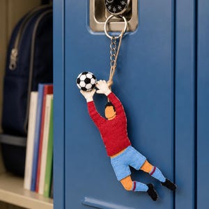 May include: A soccer-themed keychain featuring a felt goalkeeper in a red shirt and blue shorts, reaching for a black and white soccer ball. The keychain hangs from a blue locker with a combination lock.