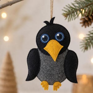 May include: A handmade felt crow ornament with a black body, gray chest, and yellow beak and feet. The crow has large blue eyes with black pupils and is hanging from a natural twine string. The ornament is suspended from a tree branch.