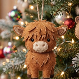May include: A brown felt Highland cow Christmas ornament with a light brown muzzle and ears, and dark brown stitching. The ornament hangs from a natural twine string against a backdrop of a decorated Christmas tree with colorful ornaments and lights.