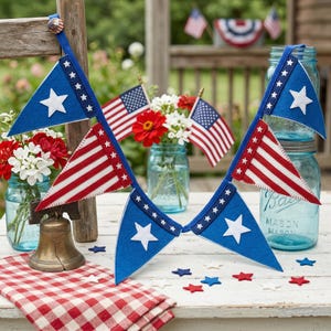 May include: A patriotic display featuring blue and red pennant flags with white stars and stripes, alongside miniature American flags and floral arrangements in mason jars. A small brass bell and star-shaped confetti complete the festive scene.