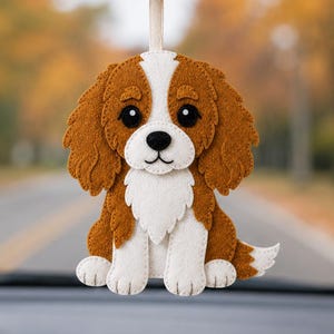 May include: A felt Cavalier King Charles Spaniel car charm. The dog has brown and white fur, with large black eyes and a black nose. It is hanging from a rearview mirror in a car, with a blurred background of a road and trees.