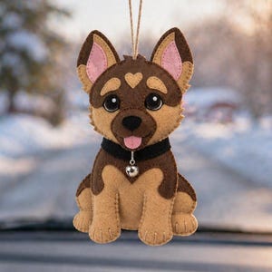 Può includere: Un ornamento per auto in feltro a forma di pastore tedesco. L'ornamento è marrone, beige e nero con l'interno delle orecchie rosa e un collare nero con una campanella argentata. È appeso a un filo davanti al cruscotto e allo specchietto retrovisore di un'auto.
