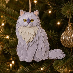 May include: A layered felt cat ornament in shades of light purple and cream, featuring bright yellow eyes and a small nose. The cat is depicted in a seated position with a fluffy chest and tail, suspended by a thin brown string from a branch.