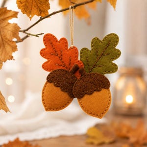 May include: A felt hanging ornament featuring two acorns with dark brown caps and light brown bodies, accompanied by one orange oak leaf and one olive green oak leaf. The pieces are joined with visible stitching and suspended by a thin twine loop.