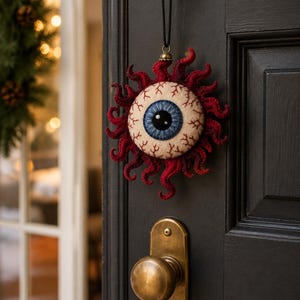 Könnte beinhalten: Ein rundes, gefilztes Hängeornament in Form eines menschlichen Augapfels mit blauer Iris, schwarzer Pupille und weißer Lederhaut mit roten gestickten Adern. Der Augapfel ist von einem Kranz aus dunkelroten, wellenförmigen Filzfasern umgeben.