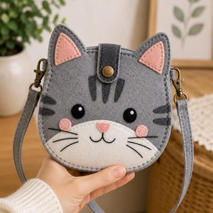 Pode incluir: Uma bolsa transversal de feltro cinza em forma de gato com focinho branco, interior das orelhas rosa e bochechas rosadas. A bolsa apresenta listras cinza-escuras, bigodes bordados em preto, olhos pretos e um fecho central com botão de pressão.