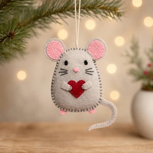 Può includere: Un ornamento in feltro grigio a forma di topo con orecchie interne rosa, occhi e baffi ricamati in nero e un piccolo naso rosa. Il topo tiene un cuore in feltro rosso tra le zampe anteriori e presenta una lunga coda grigia ricurva.