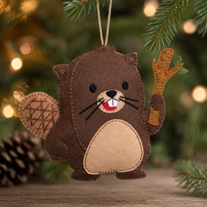 May include: A felt beaver ornament featuring a dark brown body with a light tan belly, a textured tail, and a small brown stick held in one paw. The beaver has black eyes, black whiskers, and a red mouth with two white teeth, suspended by a string.