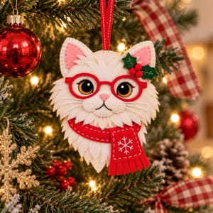 May include: A felt cat ornament featuring a white face with large brown eyes, wearing bright red glasses and a red scarf with a white snowflake pattern. The cat has pink inner ears and a small green holly sprig with red berries attached to one ear.