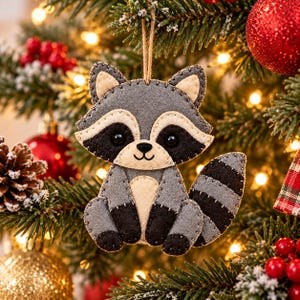 May include: A felt raccoon ornament featuring a grey body, cream-colored face and belly, and black eye patches, nose, and tail stripes. The figure is constructed with visible dark stitching along the edges and hangs from a thin brown twine loop.