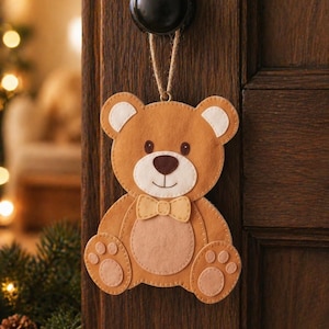 May include: A brown felt teddy bear door hanger with a light brown bow tie and light brown paw pads. The bear has white inner ears and a brown nose. It hangs from a black door knob with a natural fiber string.