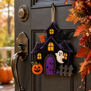May include: A black felt haunted house wall hanging featuring a purple door, orange pumpkin, white ghost, and yellow windows. The house has a brick chimney, a small gray fence, and a black bat silhouette, all suspended by a jute twine loop.