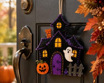 May include: A felt hanging decoration shaped like a haunted house, featuring a black structure with a purple door, yellow windows, an orange chimney, a white ghost, a jack-o'-lantern, a grey fence, and a black bat, suspended by a jute twine loop.